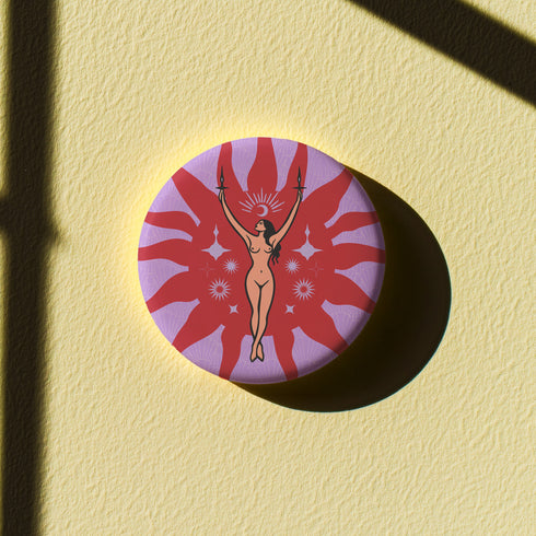 Circular pin featuring a nude goddess figure in gold against a red sunburst pattern on lavender background, with moon and star details