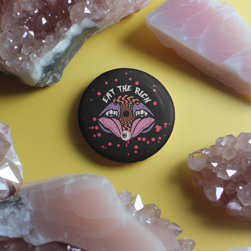 Black circular pin badge with purple fanged mouths design, psychedelic orange spiral, and "EAT THE RICH" text in white, scattered with red dots