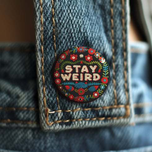 Stay Weird" black pinback button with pearl-effect lettering surrounded by folk art flowers in red and turquoise on a dark background.
