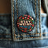 Stay Weird" black pinback button with pearl-effect lettering surrounded by folk art flowers in red and turquoise on a dark background.