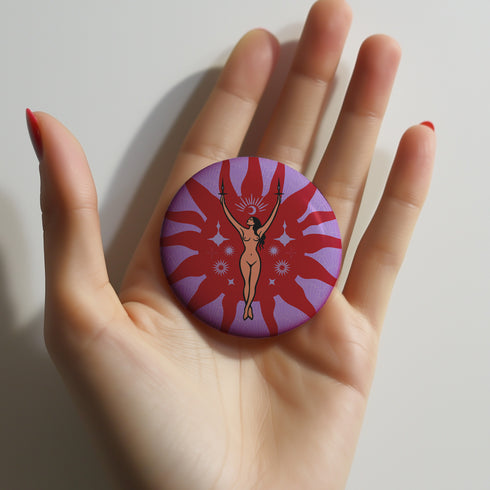 Circular pin featuring a nude goddess figure in gold against a red sunburst pattern on lavender background, with moon and star details