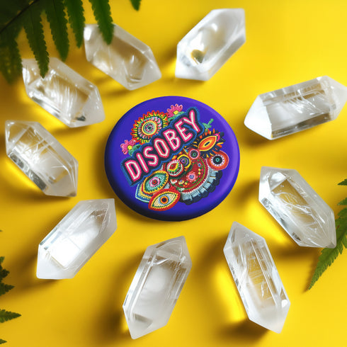 Stay Weird" black pinback button with pearl-effect lettering surrounded by folk art flowers in red and turquoise on a dark background.