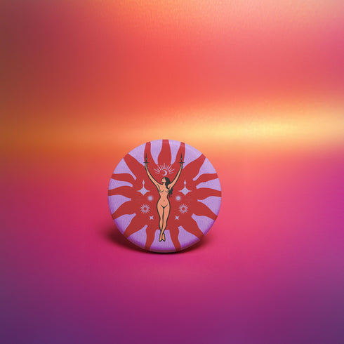 Circular pin featuring a nude goddess figure in gold against a red sunburst pattern on lavender background, with moon and star details