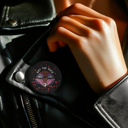 Black circular pin badge with purple fanged mouths design, psychedelic orange spiral, and "EAT THE RICH" text in white, scattered with red dots