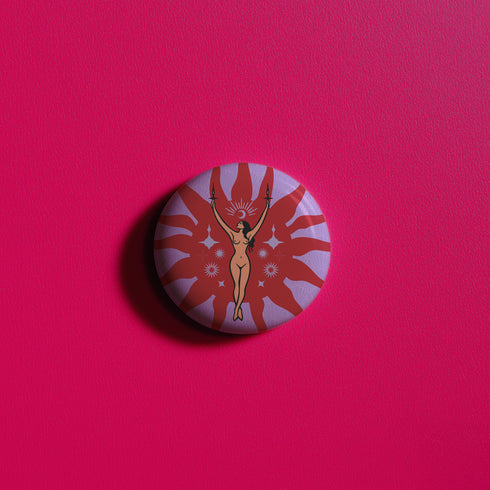 Circular pin featuring a nude goddess figure in gold against a red sunburst pattern on lavender background, with moon and star details