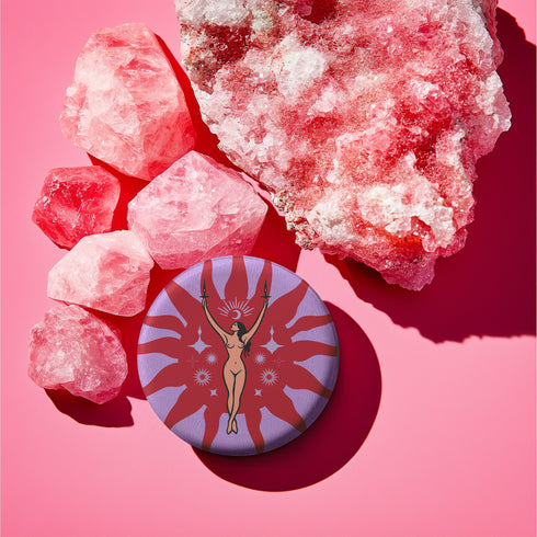 Circular pin featuring a nude goddess figure in gold against a red sunburst pattern on lavender background, with moon and star details