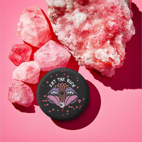 Black circular pin badge with purple fanged mouths design, psychedelic orange spiral, and "EAT THE RICH" text in white, scattered with red dots