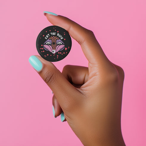 Black circular pin badge with purple fanged mouths design, psychedelic orange spiral, and "EAT THE RICH" text in white, scattered with red dots