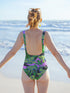 A one-piece scoop neck swimsuit featuring a bold, all-over scattered print. The flat, 2D printed design has a solid black background overlaid with bright pink abstract shapes, medium green tropical leaves, and striking lime green tigers with dark navy blue stripes. The bathing suit features a low scoop back, cheeky fit bottom, wide shoulder straps, and a double-layer front with white lining inside. The fabric has zig-zag stitching along the edges.