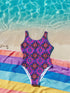 Bold patterned one-piece swimsuit with retro evil eye floral design, styled in festival-ready outdoor settings. Swimsuit features a scoop neckline, cheeky fit, and low scoop back. Ideal for summer vacations, pool parties, raves, and alt girl fashion. Made from stretchy, moisture-wicking fabric with UPF 50+ protection. Available in sizes XS to 3X. Colorful, statement swimwear designed for confident, maximalist style lovers.