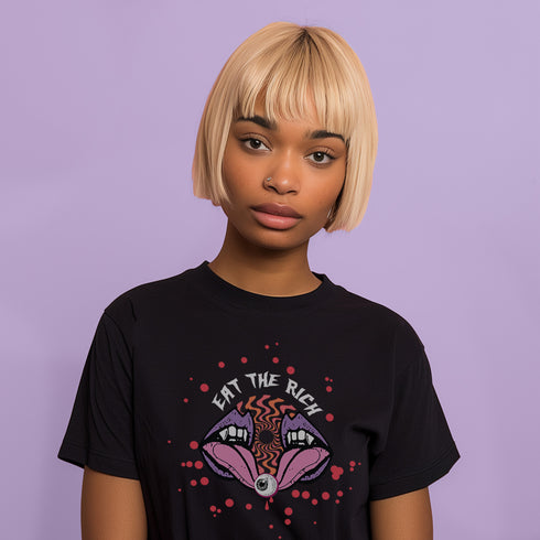 White t-shirt featuring a surreal, psychedelic design centered on the chest. An open mouth with sharp vampire-like teeth and pink tongue dominates the image. Between the teeth, a hypnotic swirling pattern in rainbow colors leads to a single eyeball on the tongue. "EAT THE RICH" is written in white above the mouth. Red paint-like splatters surround the entire design, giving it a grungy, punk aesthetic. The shirt is from the "Honey Dazed" brand.