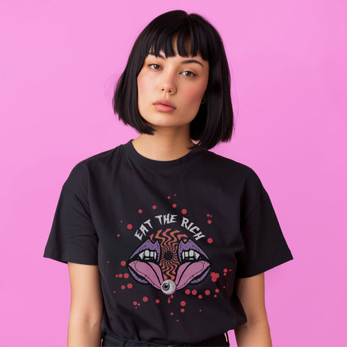 White t-shirt featuring a surreal, psychedelic design centered on the chest. An open mouth with sharp vampire-like teeth and pink tongue dominates the image. Between the teeth, a hypnotic swirling pattern in rainbow colors leads to a single eyeball on the tongue. "EAT THE RICH" is written in white above the mouth. Red paint-like splatters surround the entire design, giving it a grungy, punk aesthetic. The shirt is from the "Honey Dazed" brand.