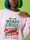 A holiday sweatshirt in pink or red with a punk and goth aesthetic. The text reads "MERRY CRISIS" and "HAPPY NEW FEAR" in a bold, gothic-style font. The design is decorated with holly leaves, berries, thorny vines, and safety pins, all arranged in a festive but alternative holiday style. This funny goth Christmas pullover is a sarcastic statement piece, perfect as dark humor apparel for the winter season.
