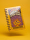 A glossy 5x7 inch spiral bound notebook featuring the phrase Inner Peace Outer Chaos in retro 70s typography. The cover showcases a vibrant psychedelic design on a mustard yellow background with a central trippy sun motif containing three mystical eyes. Below the sun, text reads Meditations, Mantras, and Meltdown Notes in bold black font. The journal has a durable black metal spiral binding and contains 150 lined pages for writing, shadow work, or daily planning.