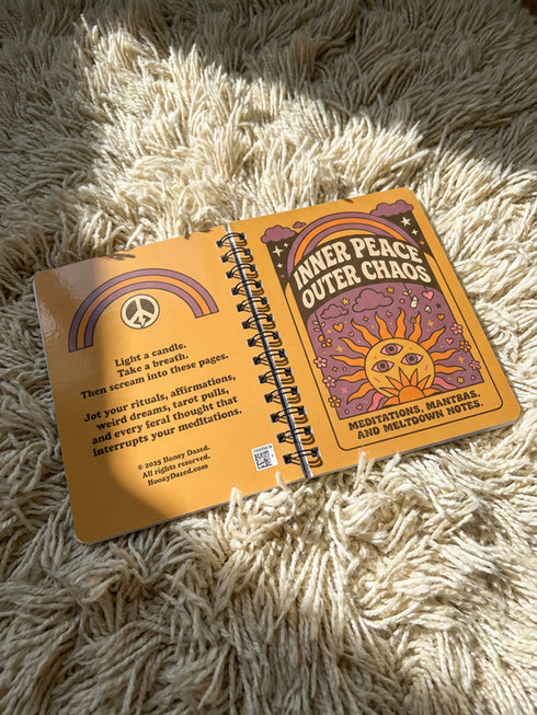 A glossy 5x7 inch spiral bound notebook featuring the phrase Inner Peace Outer Chaos in retro 70s typography. The cover showcases a vibrant psychedelic design on a mustard yellow background with a central trippy sun motif containing three mystical eyes. Below the sun, text reads Meditations, Mantras, and Meltdown Notes in bold black font. The journal has a durable black metal spiral binding and contains 150 lined pages for writing, shadow work, or daily planning.