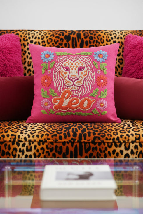 Maximalist throw pillow for the Leo zodiac sign featuring a centralized lion head medallion with an ornate mane mimicking embroidered textures. The background is a vibrant hot pink, accented by ochre, rust, and lime green floral motifs and blue flowers in each corner. The pillow is square with a hidden zipper and a polyester insert. This high-contrast design is a perfect fit for dopamine decor, whimsigoth apartments, or any eclectic home looking for a bold astrology-inspired statement piece.