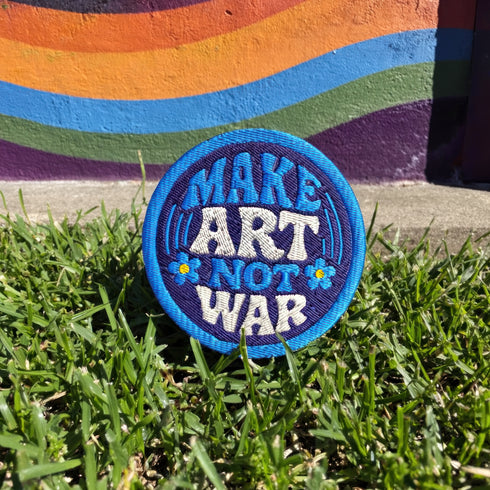 Round embroidered iron-on patch featuring "MAKE ART NOT WAR" text with blue floral design. Navy blue background with light blue border, white cream lettering, and small yellow-centered blue flowers. 3-inch circular twill fabric patch perfect for jackets, backpacks, denim, and clothing customization. Anti-war peace activist feminist punk alternative accessory for creative counterculture gift giving.