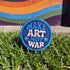 Round embroidered iron-on patch featuring "MAKE ART NOT WAR" text with blue floral design. Navy blue background with light blue border, white cream lettering, and small yellow-centered blue flowers. 3-inch circular twill fabric patch perfect for jackets, backpacks, denim, and clothing customization. Anti-war peace activist feminist punk alternative accessory for creative counterculture gift giving.