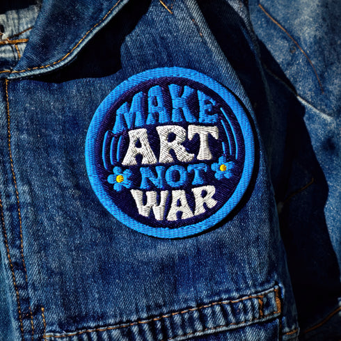 Round embroidered iron-on patch featuring "MAKE ART NOT WAR" text with blue floral design. Navy blue background with light blue border, white cream lettering, and small yellow-centered blue flowers. 3-inch circular twill fabric patch perfect for jackets, backpacks, denim, and clothing customization. Anti-war peace activist feminist punk alternative accessory for creative counterculture gift giving.