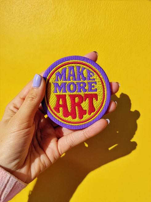 A round embroidered patch with the words "MAKE MORE ART" in a retro 70s psychedelic font. The word "ART" is magenta pink, "MAKE MORE" is purple, and they are set against a bright yellow circular background. The entire design is encircled by a thin pink line and a thick purple border. This 3-inch iron-on applique is a colorful, groovy accessory for DIY projects, perfect for personalizing jackets, backpacks, or denim vests. It serves as an inspirational artist gift with a vintage aesthetic.