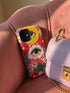 Maximalist phone case with a vibrant hot pink background featuring a faux crochet knit texture illusion. The design includes a large yellow crescent moon with a green eye in the top right, a central white cloud with a green eye and red lashes, and green mountain peaks at the bottom. Red flowers and pink swirling mist accent the scene.