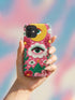 Maximalist phone case with a vibrant hot pink background featuring a faux crochet knit texture illusion. The design includes a large yellow crescent moon with a green eye in the top right, a central white cloud with a green eye and red lashes, and green mountain peaks at the bottom. Red flowers and pink swirling mist accent the scene.
