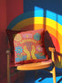 A square red decorative pillow featuring a flat, faux-embroidery design of the Aquarius zodiac sign. The word "AQUARIUS" curves in yellow letters above two yellow hands catching falling blue raindrops. Stylized pink and blue folk-art flowers and green leaves flank the hands, with blue waves at the bottom. The design mimics the texture of stitched thread but is a smooth 2D print.