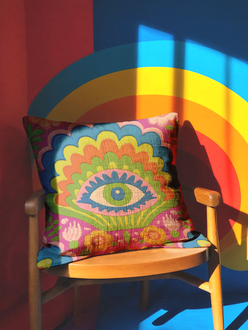 Polyester faux embroidered pillow cover featuring a bold maximalist evil eye design with a colorful folk-art inspired pattern. The front print includes a central blue, green, and pink eye surrounded by scalloped floral shapes, rainbow-like arches, leafy details, and bright decorative flowers in pink, orange, yellow, blue, and green. Soft square pillow cover with a textured woven-look print. Available in 18x18 or 22x22 inches.