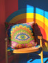 Polyester faux embroidered pillow cover featuring a bold maximalist evil eye design with a colorful folk-art inspired pattern. The front print includes a central blue, green, and pink eye surrounded by scalloped floral shapes, rainbow-like arches, leafy details, and bright decorative flowers in pink, orange, yellow, blue, and green. Soft square pillow cover with a textured woven-look print. Available in 18x18 or 22x22 inches.