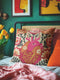 A square decorative pillow cover made of beige faux-linen fabric featuring a large, colorful illustration of an anatomical heart in the center. The heart is depicted in shades of bright pink, red, and orange with gold veins. Stylized flowers and botanical vines sprout from the arteries of the heart, featuring orange hibiscus-style blooms, red petals, and dark green leafy stems. The design mimics the look of textured embroidery but is a smooth flat print.