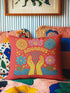 A square red decorative pillow featuring a flat, faux-embroidery design of the Aquarius zodiac sign. The word "AQUARIUS" curves in yellow letters above two yellow hands catching falling blue raindrops. Stylized pink and blue folk-art flowers and green leaves flank the hands, with blue waves at the bottom. The design mimics the texture of stitched thread but is a smooth 2D print.