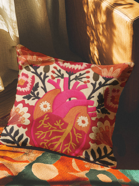 A square decorative pillow cover made of beige faux-linen fabric featuring a large, colorful illustration of an anatomical heart in the center. The heart is depicted in shades of bright pink, red, and orange with gold veins. Stylized flowers and botanical vines sprout from the arteries of the heart, featuring orange hibiscus-style blooms, red petals, and dark green leafy stems. The design mimics the look of textured embroidery but is a smooth flat print.