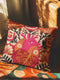 A square decorative pillow cover made of beige faux-linen fabric featuring a large, colorful illustration of an anatomical heart in the center. The heart is depicted in shades of bright pink, red, and orange with gold veins. Stylized flowers and botanical vines sprout from the arteries of the heart, featuring orange hibiscus-style blooms, red petals, and dark green leafy stems. The design mimics the look of textured embroidery but is a smooth flat print.