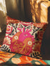 A square decorative pillow cover made of beige faux-linen fabric featuring a large, colorful illustration of an anatomical heart in the center. The heart is depicted in shades of bright pink, red, and orange with gold veins. Stylized flowers and botanical vines sprout from the arteries of the heart, featuring orange hibiscus-style blooms, red petals, and dark green leafy stems. The design mimics the look of textured embroidery but is a smooth flat print.