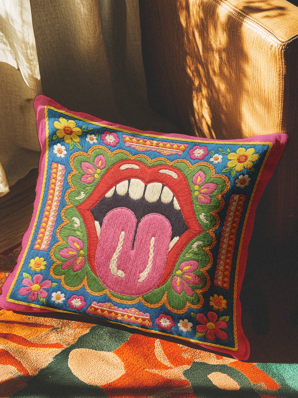 Maximalist throw pillow cover featuring a printed faux embroidery design with red open lips, pink tongue, white teeth, green scalloped medallion border, yellow and pink flowers, and decorative folk-inspired bands on a bright blue background with hot pink edging. Square colorful pillowcase for eclectic home decor, retro interiors, dopamine decor, boho living rooms, statement sofas, beds, chairs, and reading nooks.