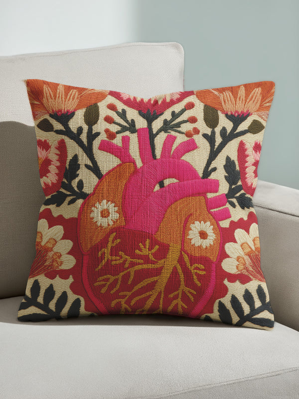 A square decorative pillow cover made of beige faux-linen fabric featuring a large, colorful illustration of an anatomical heart in the center. The heart is depicted in shades of bright pink, red, and orange with gold veins. Stylized flowers and botanical vines sprout from the arteries of the heart, featuring orange hibiscus-style blooms, red petals, and dark green leafy stems. The design mimics the look of textured embroidery but is a smooth flat print.