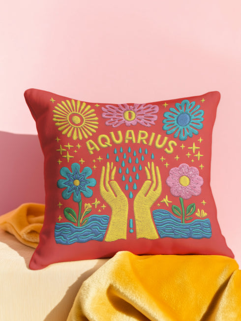 A square red decorative pillow featuring a flat, faux-embroidery design of the Aquarius zodiac sign. The word "AQUARIUS" curves in yellow letters above two yellow hands catching falling blue raindrops. Stylized pink and blue folk-art flowers and green leaves flank the hands, with blue waves at the bottom. The design mimics the texture of stitched thread but is a smooth 2D print.