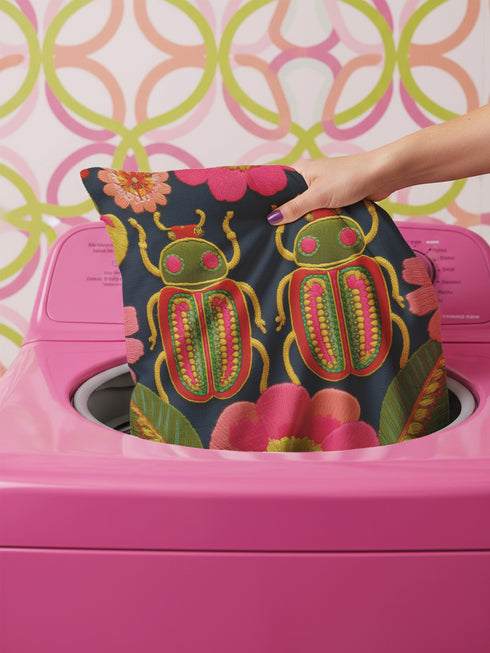 Navy blue square throw pillow cover featuring two vibrant beetles in a symmetrical design. The beetles have jewel-toned wings in emerald green and hot pink, rendered in a faux-embroidery texture style that mimics stitched threads. The background is a deep midnight blue with stylized floral accents. The texture is a smooth digital print on spun polyester. Ideal for maximalist homes and entomolgy art.