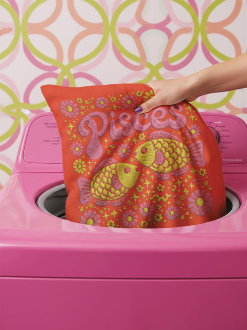 A vibrant tomato red square throw pillow featuring a retro 70s Pisces zodiac design in a faux embroidery print style. The word Pisces appears in bubblegum pink bubble letters at the top, sitting above two yellow and pink fish swimming in a circle representing the water sign. The background is densely filled with yellow and pink daisies. The texture visually mimics stitched needlework but is a smooth 2D print on linen-feel fabric. Perfect for maximalist, eclectic, and dopamine decor styles.