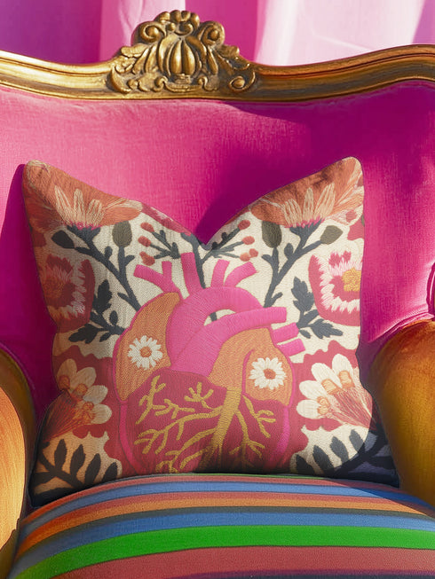 A square decorative pillow cover made of beige faux-linen fabric featuring a large, colorful illustration of an anatomical heart in the center. The heart is depicted in shades of bright pink, red, and orange with gold veins. Stylized flowers and botanical vines sprout from the arteries of the heart, featuring orange hibiscus-style blooms, red petals, and dark green leafy stems. The design mimics the look of textured embroidery but is a smooth flat print.