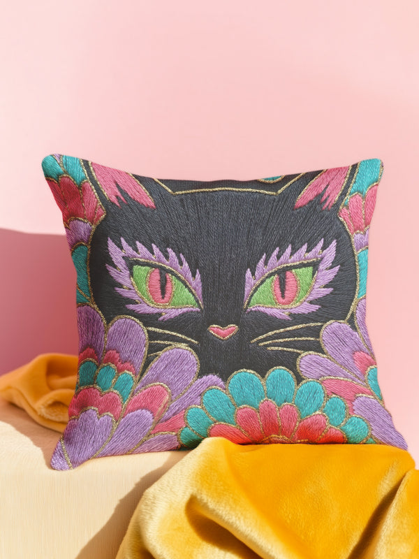 A square decorative throw pillow cover featuring a vibrant, maximalist flat print design of a black cat face. The cat has striking green eyes with pink centers and is surrounded by layered, scalloped floral and feather-like patterns in bright magenta, teal, and purple. The design includes faux gold thread outlines, giving the illusion of intricate embroidery on a dark background. The pillowcase has a textured linen-like finish and clean edges.