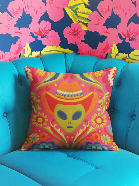 A vibrant pink square throw pillow featuring a green alien head wearing an orange cowboy hat. The alien is surrounded by colorful retro floral patterns in yellow, blue, and red. The style combines space cowgirl aesthetic with maximalist dopamine decor. The fabric has a premium linen texture. The cushion sits on a bright blue sofa, showcasing its bold, funky design. Perfect for weirdcore, sci-fi, or western themed rooms.