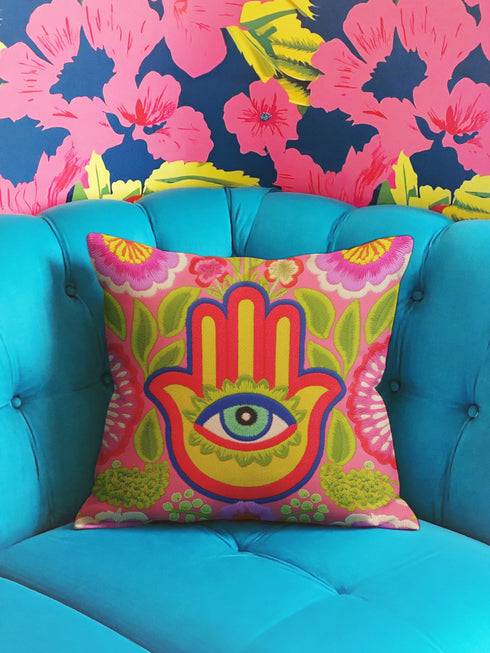 A square decorative throw pillow cover featuring a vibrant pink background with a bold, symmetrical design. In the center is a large, multi-colored Hamsa hand symbol outlined in red and yellow, containing a detailed evil eye with a green iris and black pupil. The hand is surrounded by stylized botanical elements, including pink flowers and green leaves. The design features a visual texture that mimics intricate thread embroidery but is a smooth, flat print on fabric.