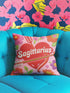 A square decorative throw pillow featuring a light pink background with a trompe-l'œil faux-embroidery print. A large red heart sits in the center, pierced by a green and white arrow. The word Sagittarius is printed across the heart in a puffy white retro font with a red drop shadow. The central design is surrounded by scattered vintage-style flowers in vibrant shades of purple, orange, and yellow with bright green leaves. The texture is a flat, high-fidelity printed illusion.
