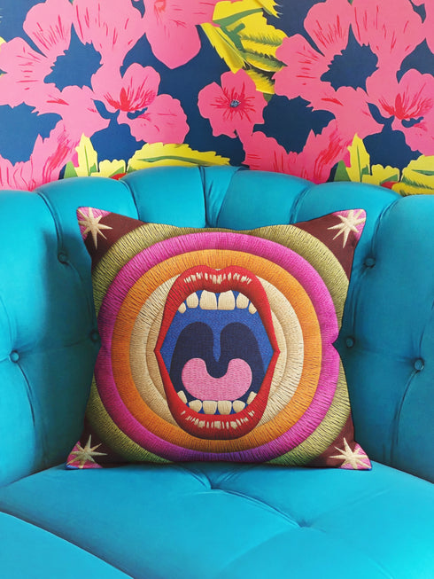 A square decorative pillowcase featuring a bold, centralized trompe-l'œil design printed to look like raised satin-stitch embroidery. The focal point is a large, open mouth with red lips, yellow teeth, and a pink tongue against a dark blue background. The mouth is surrounded by concentric radial rings in bright pink, orange, yellow, and olive green. The four corners are a dark burgundy brown, each accented with a yellow four-pointed starburst. The fabric has a woven linen-like texture.