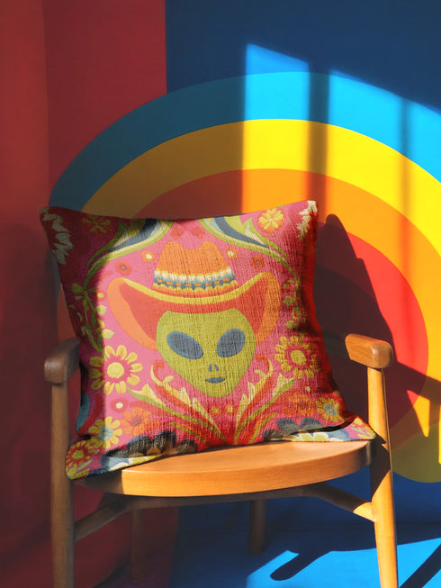 A vibrant pink square throw pillow featuring a green alien head wearing an orange cowboy hat. The alien is surrounded by colorful retro floral patterns in yellow, blue, and red. The style combines space cowgirl aesthetic with maximalist dopamine decor. The fabric has a premium linen texture. The cushion sits on a bright blue sofa, showcasing its bold, funky design. Perfect for weirdcore, sci-fi, or western themed rooms.