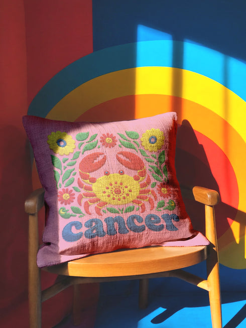 A square pink pillowcase features a vibrant, multi-colored design centered around a yellow and red crab representing the Cancer zodiac sign. The design is framed by symmetrical green leaves and yellow and red flowers. The word "cancer" is written in a bold, blue, rounded font at the bottom. The visual texture mimics intricate raised embroidery with visible stitch patterns, though the surface is a smooth 2D print on a linen-textured polyester fabric with a hidden zipper.