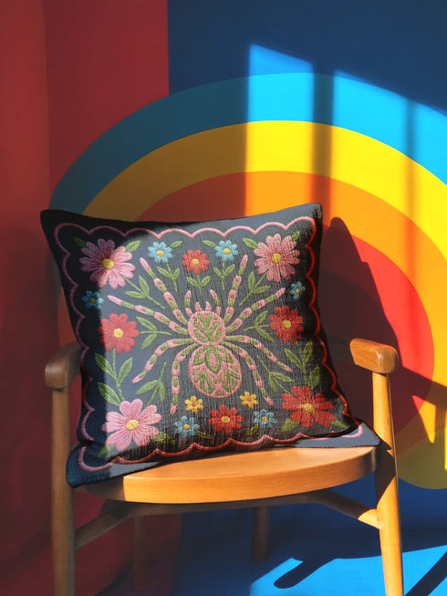 Square throw pillow cover featuring a symmetrical folk art spider illustration centered on a large hot pink flower. The spider has a striped abdomen in green, red, and yellow with pink legs. The background is a textured dark navy blue fabric with a visible faux linen weave. Surrounding the central spider are stylized botanical motifs including red daisies, blue forget-me-nots, and green leaves. The illustration is flat printed with a high-quality finish. This image shows the case only.