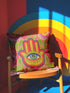A square decorative throw pillow cover featuring a vibrant pink background with a bold, symmetrical design. In the center is a large, multi-colored Hamsa hand symbol outlined in red and yellow, containing a detailed evil eye with a green iris and black pupil. The hand is surrounded by stylized botanical elements, including pink flowers and green leaves. The design features a visual texture that mimics intricate thread embroidery but is a smooth, flat print on fabric.