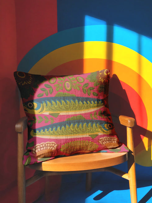 A square throw pillow cover featuring a symmetrical maximalist design on a hot pink background. Two stylized green and blue sardines swim in opposite directions in the center, featuring detailed scales and yellow eyes. The fish are surrounded by a folk art style pattern of green leafy vines and stylized orange and yellow flowers at the corners. The fabric has a faux linen texture with a flat printed design that mimics the look of vintage embroidery. Vibrant colors include pink, green, blue.